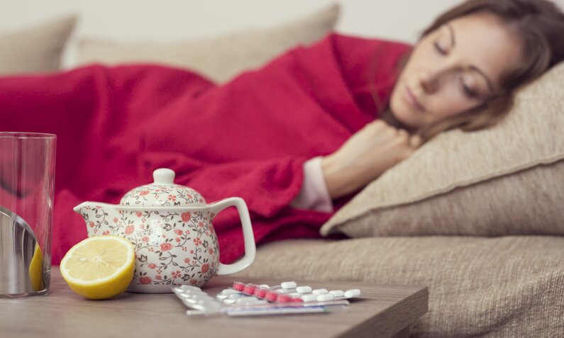 Frau liegt mit roter Decke zugedeckt auf dem Sofa, vor ihr steht eine Teekanne und eine aufgeschnittene Zitrone. | © Getty Images / vladans