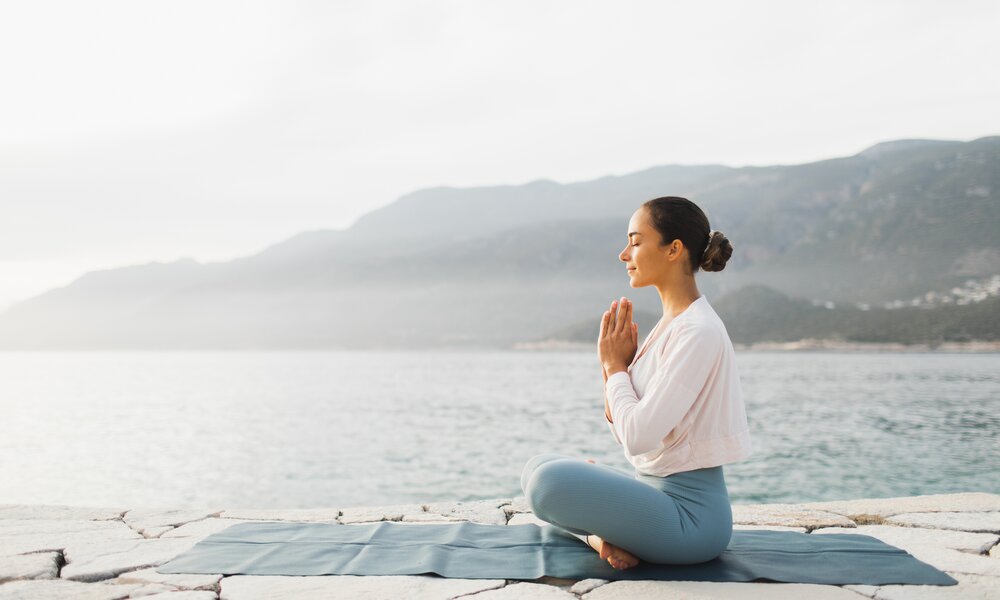 Junge Frau mit dunklem Haar sitzt vor einem See in Yoga-Pose und mit geschlossenen Augen auf einer Matte und im Schneidersitz. | © Getty Images / Oleg Breslavtsev