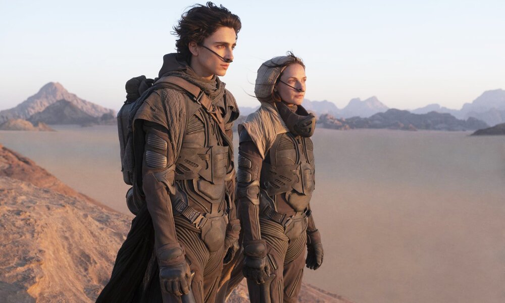 Timothy Chalamet und Rebecca Ferguson in futuristischer Kleidung vor einer Wüstenlandschaft in einer Szene des Kinofilms "Dune" | © Warner Bros.