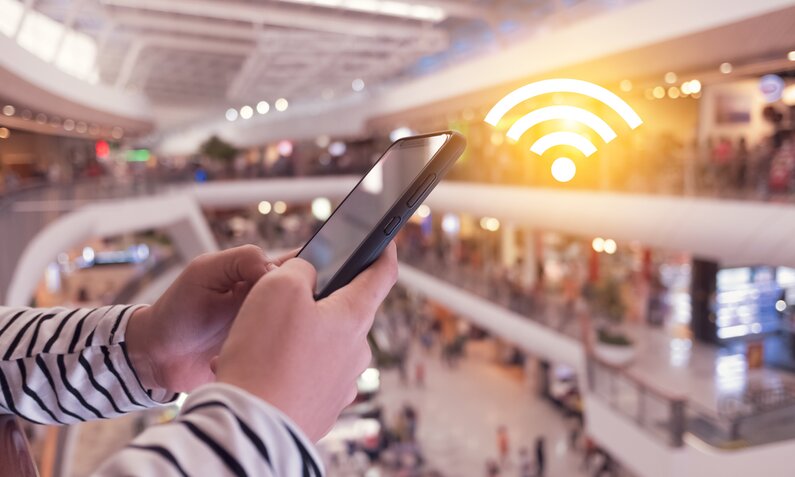 Hände halten ein Smartphone, im Hintergrund ist eine Mall zu sehen, ein WLAN-Symbol ist über dem Smartphone ins Bild kopiert. | © Getty Images/Suwaree Tangbovornpichet