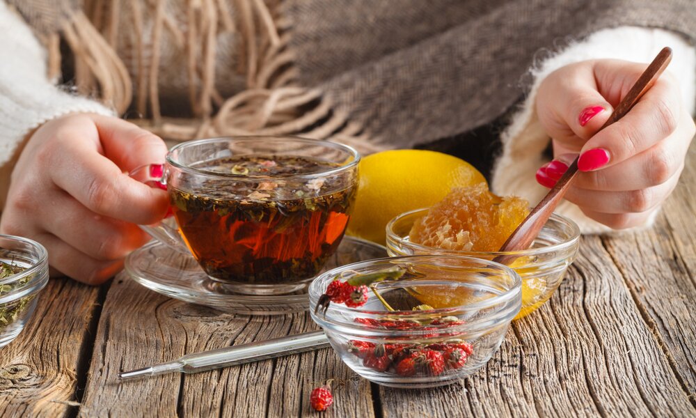 Die Hände einer jungen Frau mit rotem Nagellack halten eine durchsichtige Tasse mit einem dampfenden Getränk. Sie trägt einen Schal  und neben der Tasse befinden sich Honig und Zitrone | © Getty Images / AndreyCherkasov
