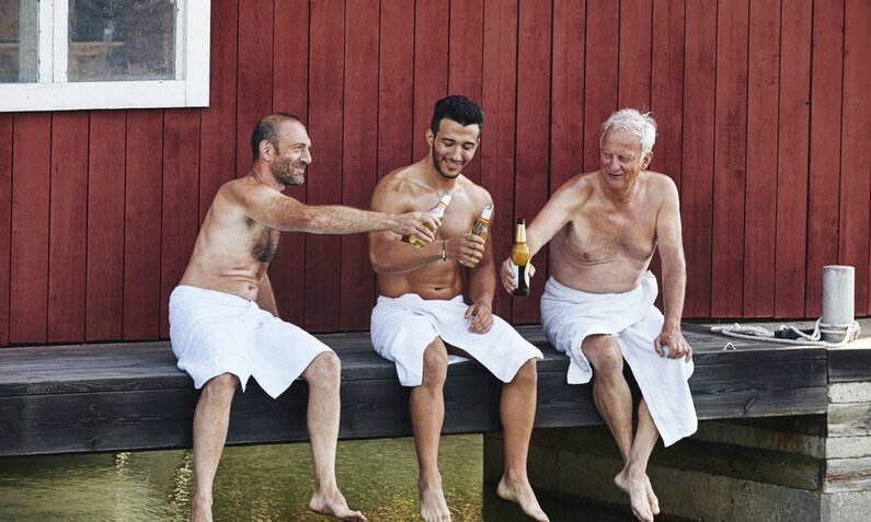 Drei Männer sitzen nach der Sauna auf einem Steg und stoßen mit Bier an  | ©  Getty Images / Frank and Helena
