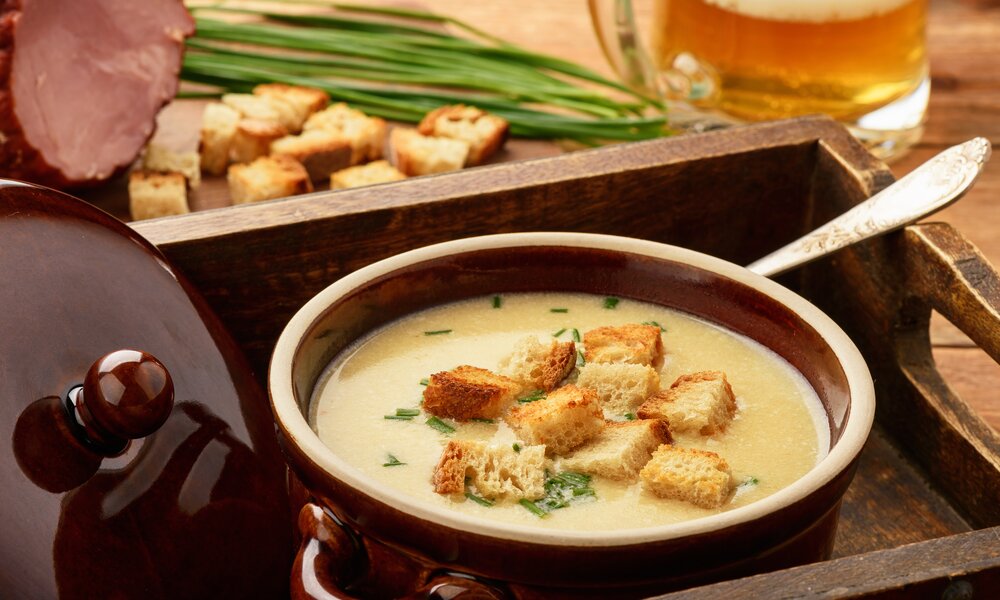 In einem Topf befindet sich eine Cremesuppe mit Croutons. Daneben steht ein Glas Bier so wie ein angeschnittener Schinken. | © Getty Images / iko636