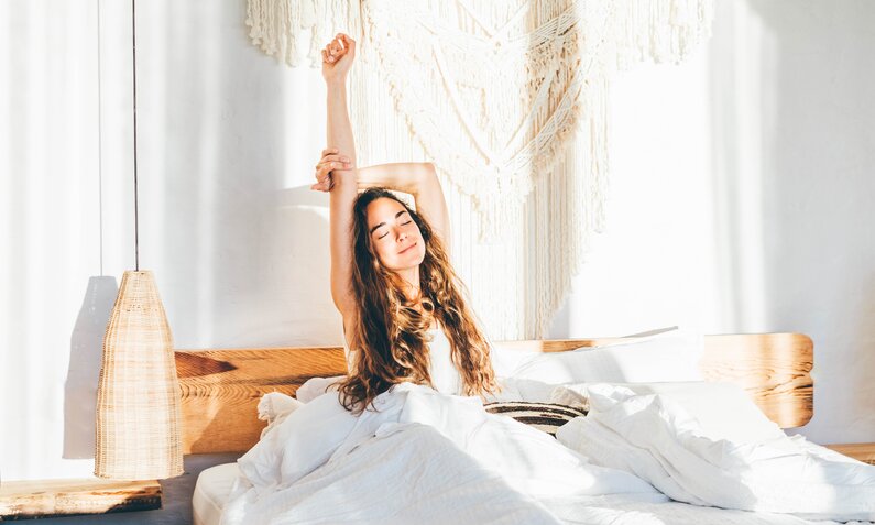 Junge Frau mit dunklen langen Haaren reckt und streckt sich im Bett. | © Getty Images / Maria Korneeva