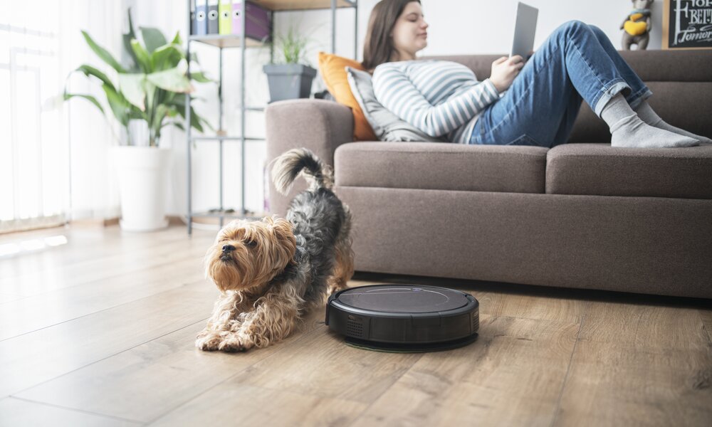 Eine junge Frau liegt in einem Wohnzimmer auf der Couch, auf dem Boden vor ihr ist ein Saugroboter unterwegs, links neben dem Gerät streckt sich ein Hund- | © Getty Images/ArtistGNDphotography