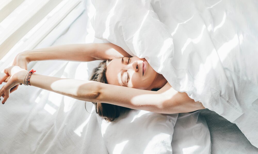 Junge Frau mit braunen Haaren liegt im Bett und streckt sich.