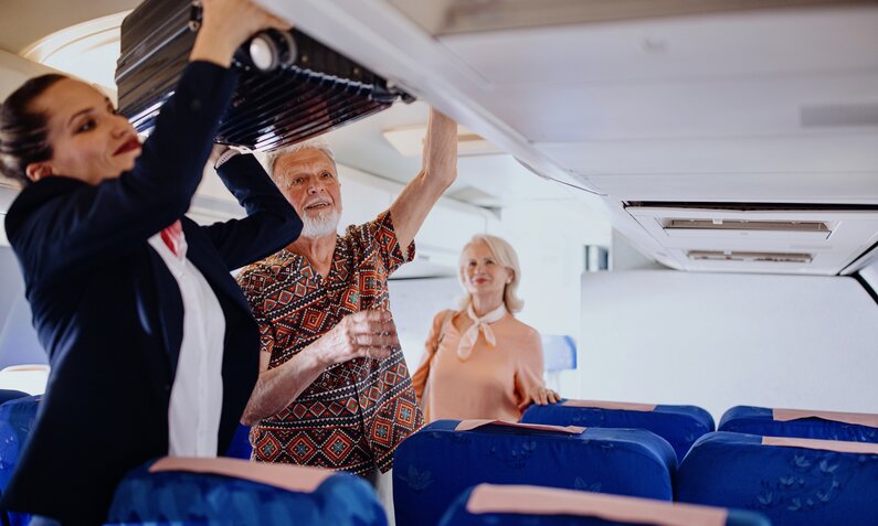 Stewardess hilft älteren Leuten beim Einräumen des Gepäcks | ©  Getty Images / Anchiy