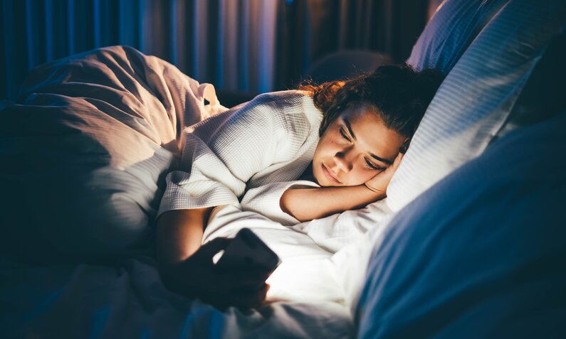 Junge Frau liegt nachts im Bett und schaut auf ihr Handy. | © Getty Images / Maria Korneeva