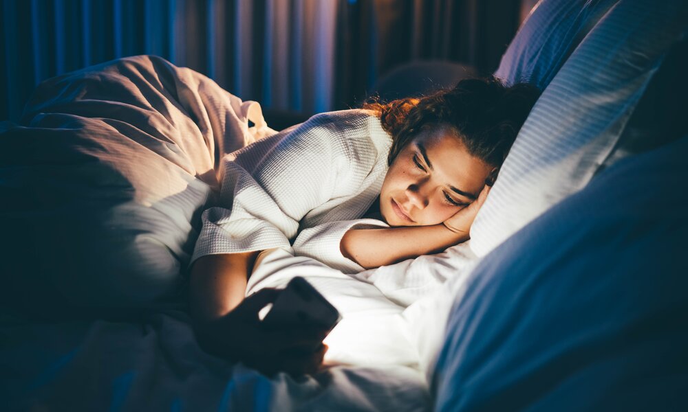 Junge Frau liegt nachts im Bett und schaut auf ihr Handy. | © Getty Images / Maria Korneeva