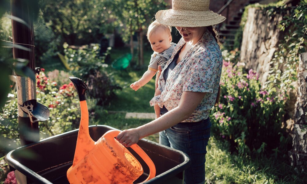 Eine junge Frau hält ein Baby auf dem Arm und tunkt eine Gießkanne in einen vollen Regenwassertank im Garten. | © Getty Images / Guido Mieth