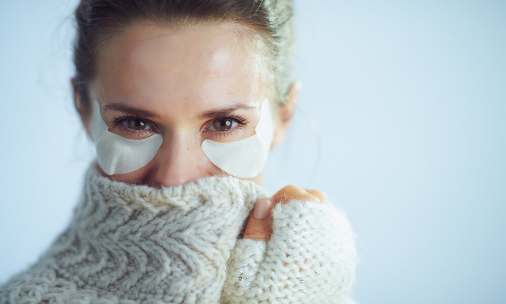 Eine Frau mittleren Alters trägt einen dicken Wollpullover und trägt zusätzlich unter den Augen feuchtigkeitsspendende Augen-Pads. | © Getty Images / CentralITAlliance