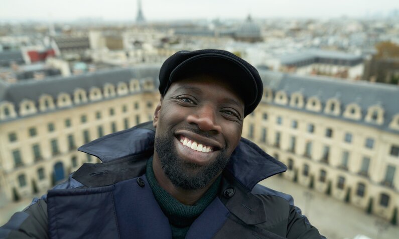 Omar Sy als Assane Diop macht ein Selfie von sich vor Pariser Häuserkulisse in einer Szene aus der Netflix-Serie "Lupin" - Staffel 3 | © Netflix