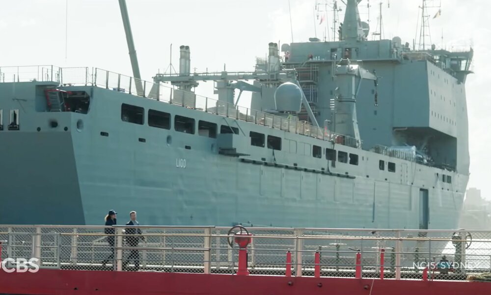 Zwei winzig wirkende Menschen laufen auf einer Brücke an einemriesigen Schiff vorbei in einer Szene der CBS-Serie "NCIS: Sydney" | © Screenshot Trailer YouTube/CBS/Paramount+