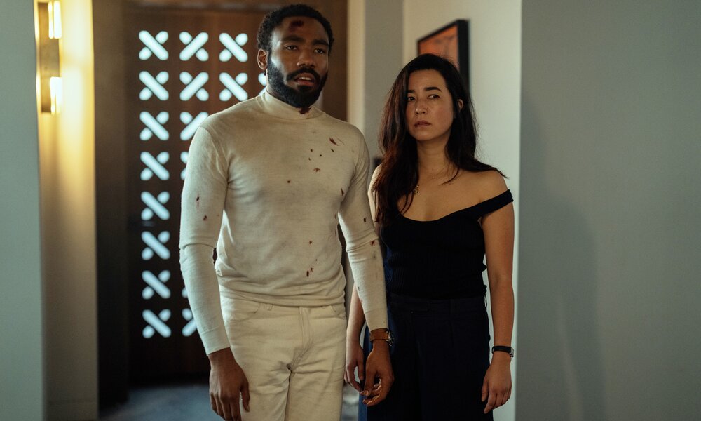 Donald Glover und Maya Erskine stehen in einer Szene aus der Prime Video Serie "Mr. & Mrs. Smith" in einem Flur, die Kleidung leicht ramponiert wie nach einem Kampf und mit ernsten Gesichtern | © Amazon Prime Video
