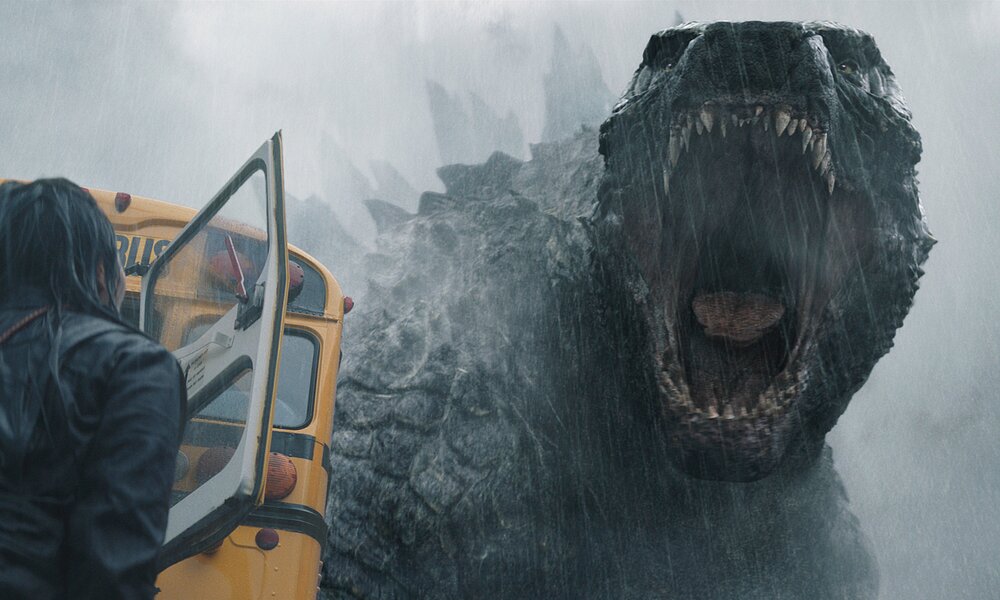 Riesiges Godzilla-Monster brüllt in Richtung einer Frau, die mit dem Rücken zur Kamera neben einem gelben Bus steht in einer Szene aus der Apple TV+ Serie "Monarch: Legacy of Monsters" | © Apple TV+