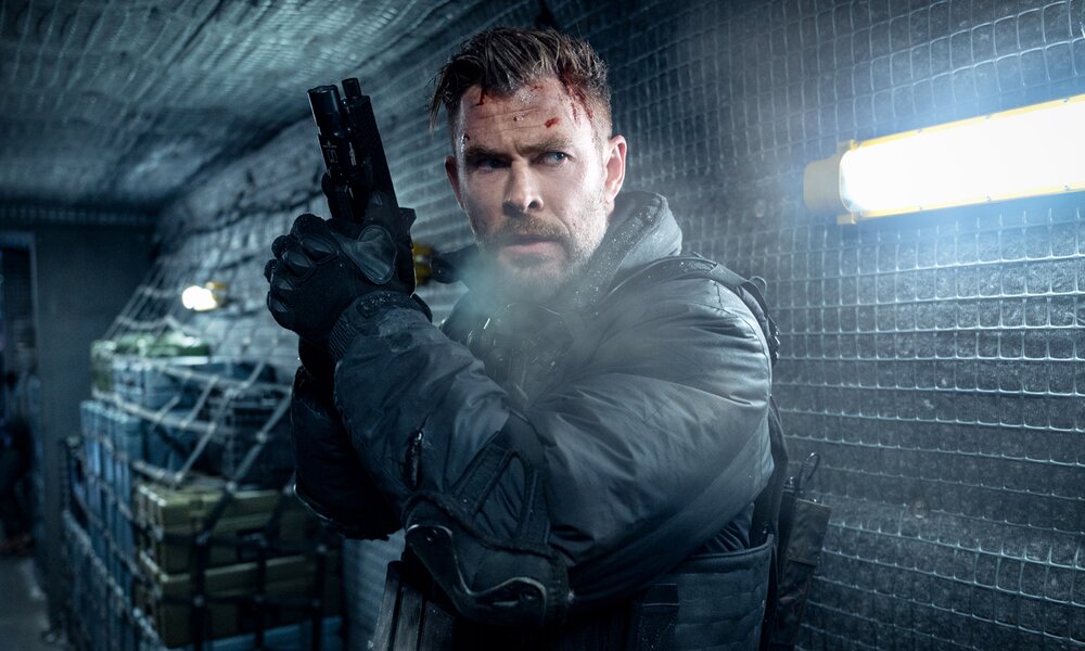 Chris Hemsworth in schwarzer Kleidung mit Waffe in einem dunklen Raum in einer Szene des Netflix-Films "Extraction 2" | © Netflix/Jasin Boland