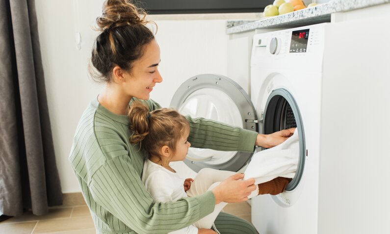 Frau mit Baby auf dem Schoss befüllt Waschmaschine mit Wäsche. | © Getty Images / Anastasiia Krivenok; 