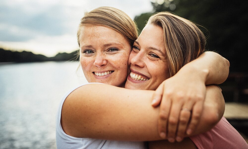 Nahaufnahme von zwei Frauen, die vor eine Naturkulisse mit See links und Wald rechts stehen. Die beiden umarmen sich fest und lächeln beide. | © Getty Images/Guido Mieth