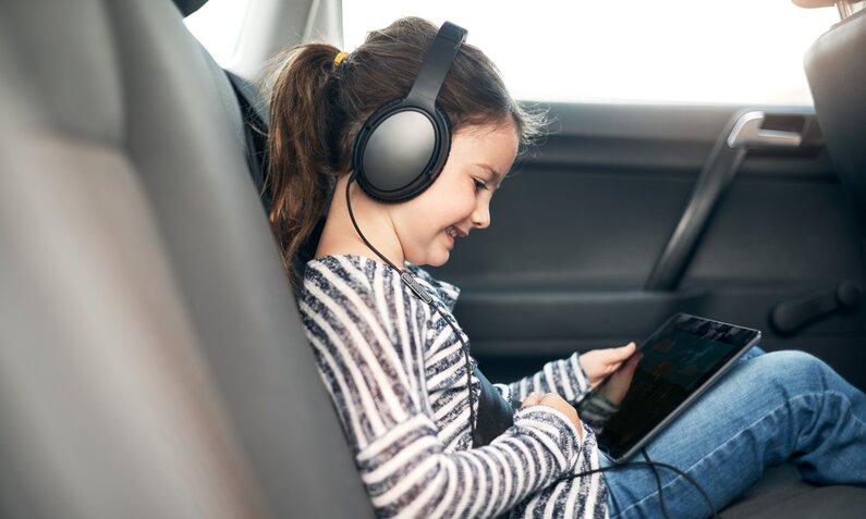 Kind sitzt mit Kopfhörern und Tablet auf der Rückbank  | ©  Getty Images / shapecharge