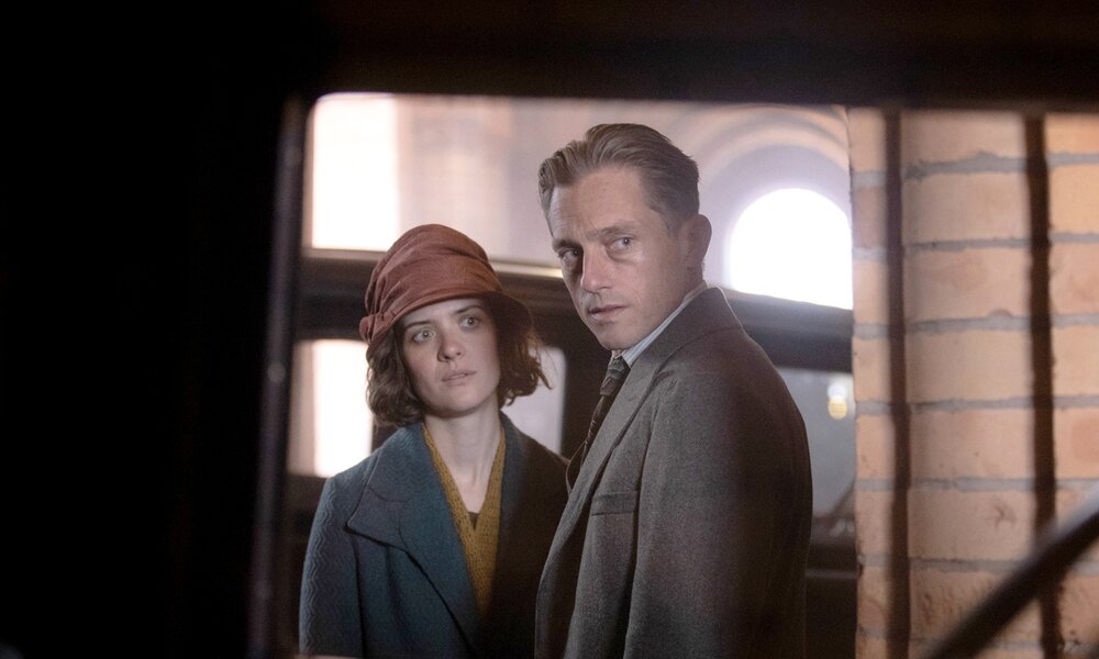 Blick aus einem Autofenster auf Liv Lisa Fries und Volker Bruch in 30er-Jahre Kleidung vor einer Backsteinmauer in einer Szene der Serie Babylon Berlin | © Frédéric Batier/X Filme Creative Pool/SKY/ARD Deget