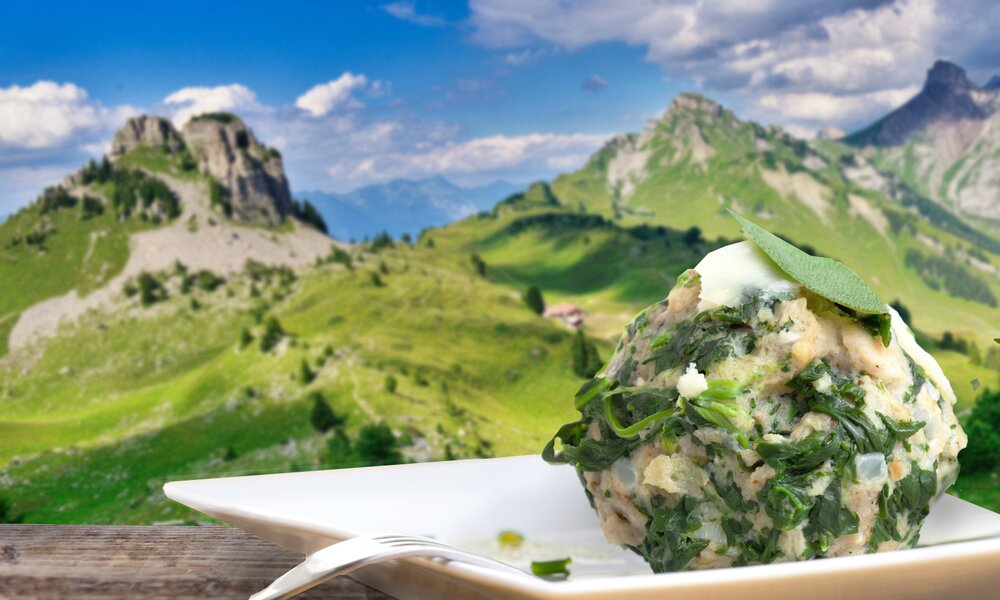 Ein köstlicher Spinatknödel auf einem weißen Teller, der auf einem rustikalen Berghütten-Tisch steht - im Hintergrund ein wunderschönes Schweizer Alpenpanorama unter blauem Himmel | © Getty Images/Zoonar RF/Pixabay