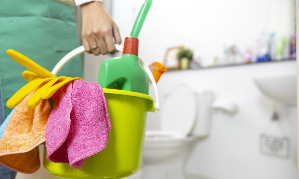 Frauenhand mit Ehering hält Putzeimmer und Putzmittel in der Hand und steht im Badezimmer. | © Getty Images / Witthaya Prasongsin