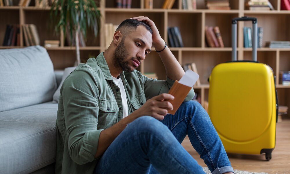 Mann sitzt enttäuscht im Wohnzimmer mit Tickets in der Hand. Im Hintergrund ein Koffer. | ©  Getty Images / Prostock-Studio