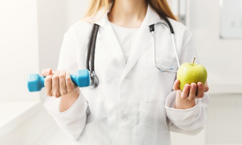 Ärztin hält Hantel und Apfel | © Getty Images / Prostock-Studio