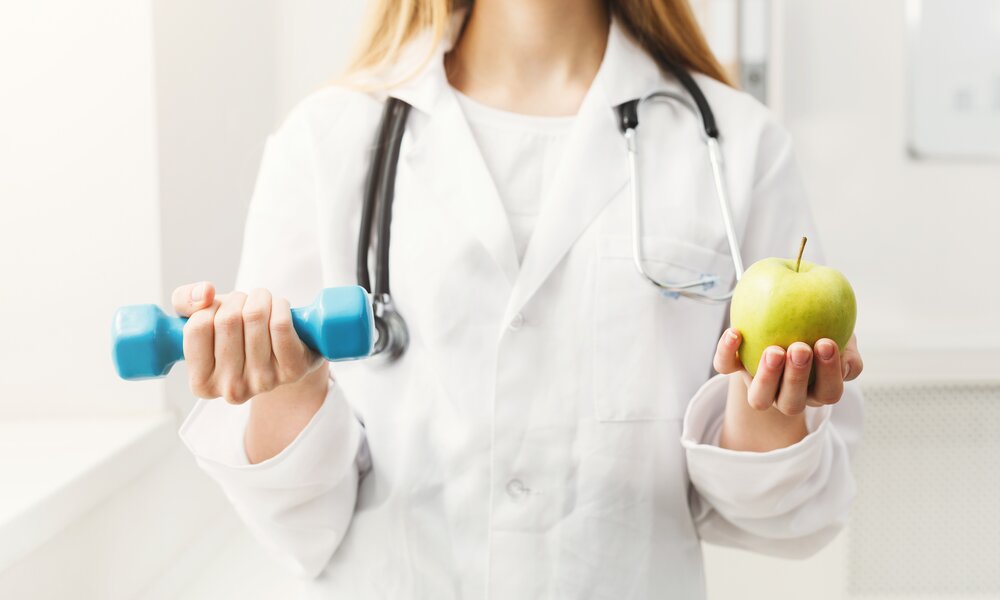 Ärztin hält Hantel und Apfel | © Getty Images / Prostock-Studio