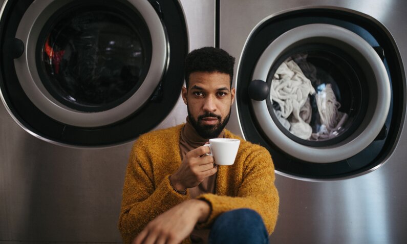 Junger Mann sitzt mit einer Tasse Kaffee vor zwei Waschmaschinen. | © Getty Images / Halfpoint Images