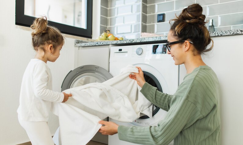 Eine junge Frau mit braunen Haaren hockt mit einem kleinen Kind vor einer Waschmaschine und packt Wäsche hinein. | © Getty Images / Anastasiia Krivenok