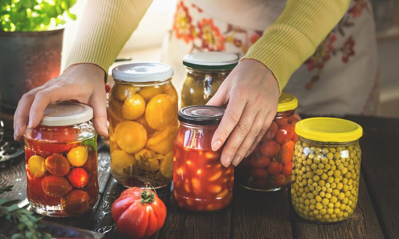 Die Hände einer Frau halten ein Einmachglas mit Lebensmitteln befüllt in den Händen. Daneben stehen weitere Einmachgläser mit Lebensmitteln. | © Getty Images / VICUSCHKA