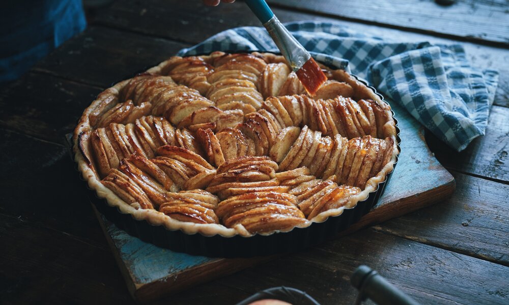 Glänzender Apfelkuchen angerichtet auf einem dunkeln Holzuntergrund. | © Getty Images / GMVozd