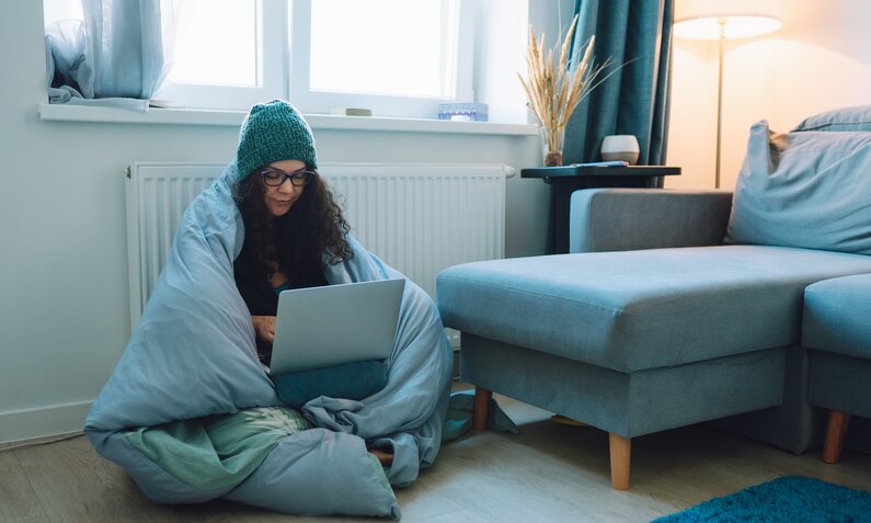 Junge Frau in Decke gehüllt sitzt vor der Heizung in einer Wohnung und schaut auf einen Laptop | © Getty Images / Olga Rolenko