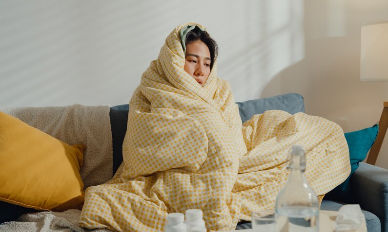 Frau sitzt erkältet eingehüllt in einen dicke Decke auf der Couch. | © Getty Images / MTStock Studio