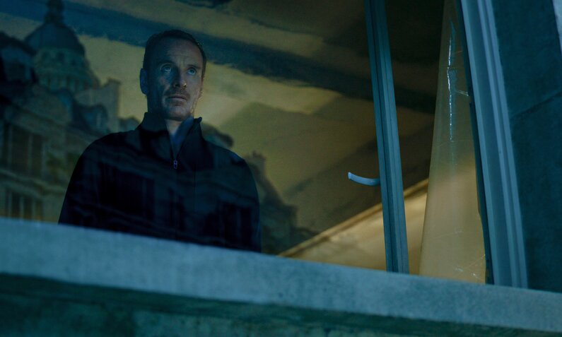 Michael Fassbender steht in einer Szene aus dem Film "The Killer" am offenen Fenster und blickt hinaus in die Nacht, hinter ihm spiiegelt sich eine Häuserzeile im Glas. | © Netflix