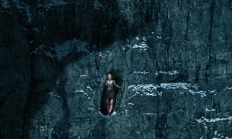Millie Bobbie Brown ist von Ferne ganz klein im Höhleneingang in einer massiven dunklen Felswand zu sehen, über die sich ein gigantischer Schatten legt - auf der Key Art zum Netflix-Film "Damsel" | © Netflix