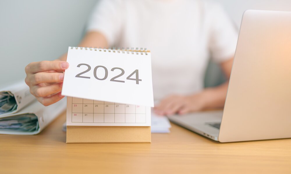 Frau sitzt an Laptop und blättert Kalender um auf das Jahr 2024 | ©  Getty Images / Panuwat Dangsungnoen