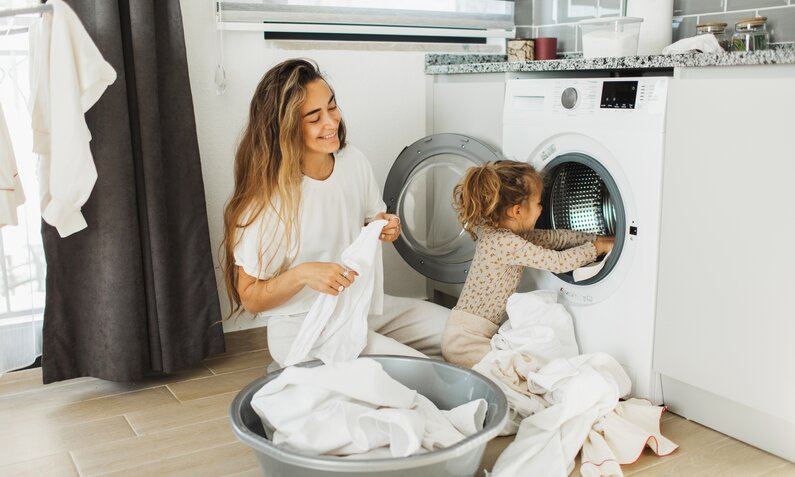 Frau und Kleinkind packen weiße Wäsche in die Waschmaschine. | © Getty Images / Anastasiia Krivenok
