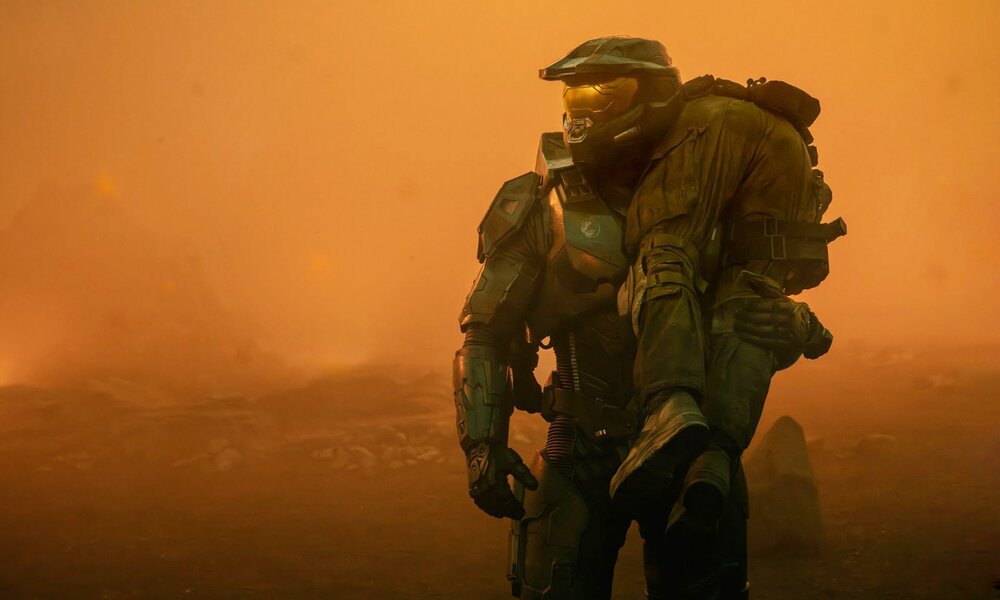 Ein Mann in einer Art Raumanzug trägt einen anderen über seiner Schulter in einer ganz in orangenes Licht getauchten Landschaft in einer Szene aus der Paramount+ Serie "Halo" | © Paramount+