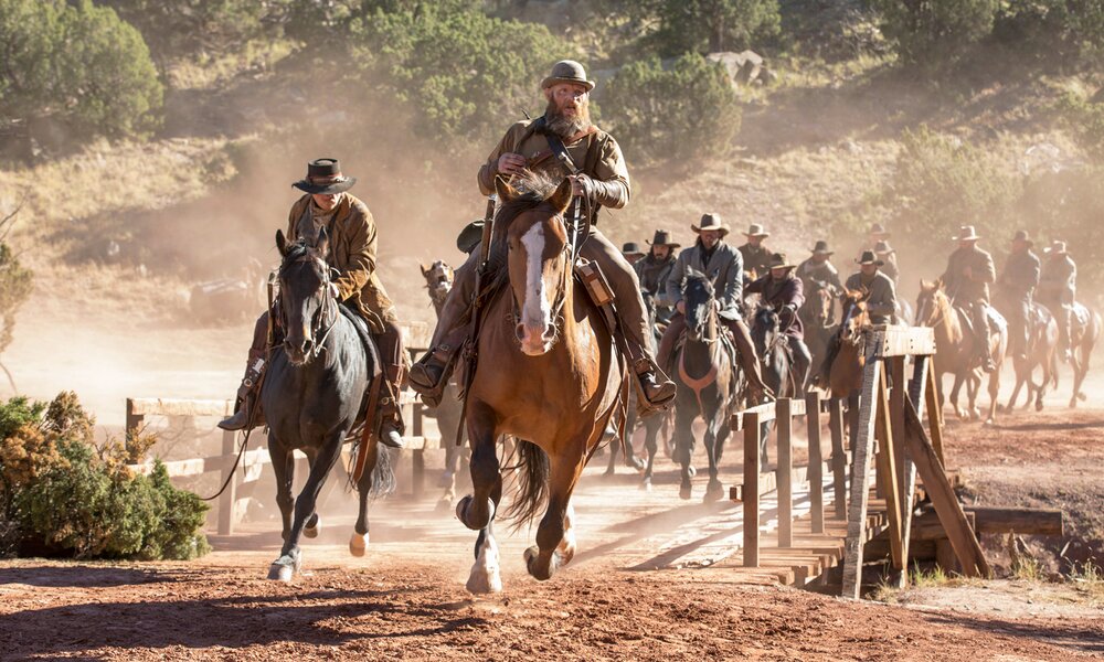 Eine Gruppe von Männern in Cowboy-Outfits reiten in vollem Galopp über eine Holzbrücke in einer Szene aus der Netflix-Serie "Godless" | © Netflix/John Britt