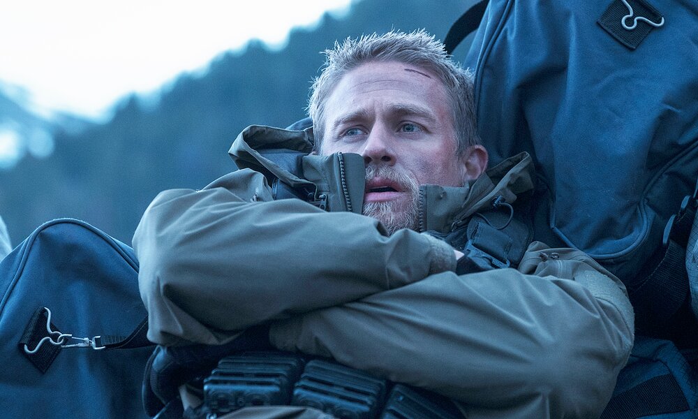 Nahaufnahme von Charlie Hunnam im armygrünen Outtfit im Freien, der sich in einer Szene aus dem Netflix-Film "Triple Frontier" mit verschränkten Armen an ein Gepäckstück lehnt | © Netflix/Melinda Sue Gordon