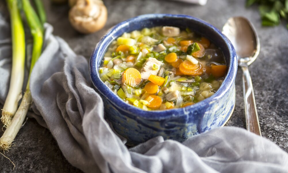 Hühnersuppe in Schale auf Holzuntergrund neben Löffel und Lauch. | © Getty Images / Westend61