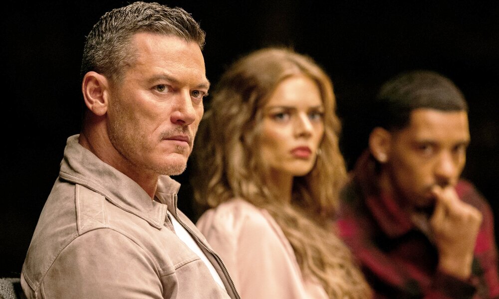 uke Evans, Samara Weaving und Melvin Gregg, sitzen  mit ernstem Gesichtsaudruck an einem Tisch vor dunklem Hintergrund in einer Szene der Serie "Nine Perfect Strangers" | © ddp images/PictureLux