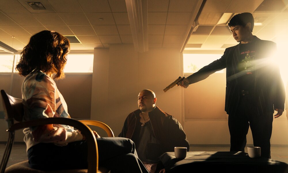 Michelle Yeoh, Jon Xue Zhang sitzen in einer Szene aus der Netflix-Serie "The Brothers Sun" in einem sonnendurchfluteten Raum, und Chau Long isteht mit gezogener Waffe vor ihnen. | © Netflix/Michael Desmond