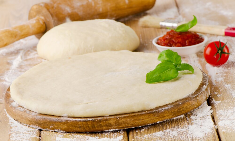 Pizzateig ausgerollt auf Holzbrett, daneben weiterer Pizzateig und Tomatensoße. | © Getty Images / bit245