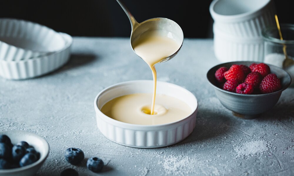 Kelle gießt Vanillesoße in kleine weiße Schüssel, daneben liegen frische Beeren. | © Getty Images / alvarez