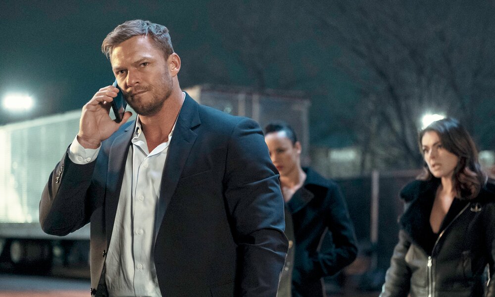 Alan Ritchson als Jack Reacher steht im weißen Hemd und mit Smartphone am Ohr im Vordergrund in einer Szene aus der Prime Video Serie "Reacher". Es ist nacht un er ist drau0en, im Hintergund sind zwi Frauuen zu sehen und ein Lastwagen