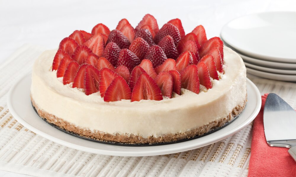 Käsekuchen drapiert mit frischen Erdbeeren auf einer Tortenplatte auf einem Platzdeckchen. | © Getty Images / 	ALEAIMAGE