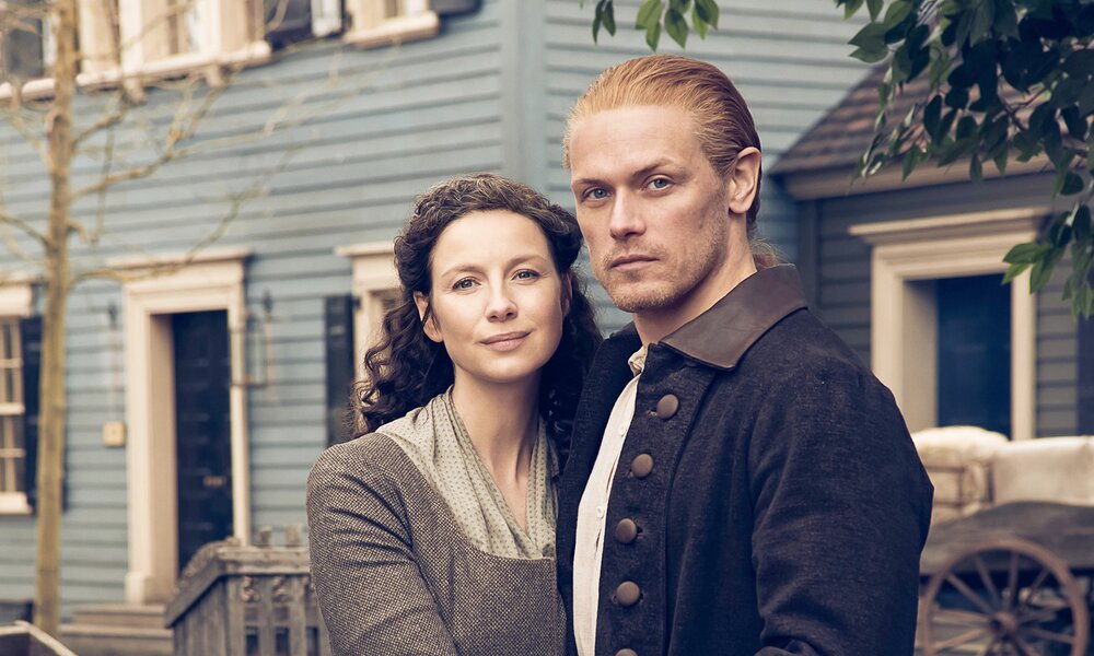 Caitriona Balfe und Sam Heughan  stehen aneinandergelehnt in historischer Kleidung vor einem Haus in einer Szene der 6. Staffel der Serie "Outlander" | © ddp images/LMKMEDIA